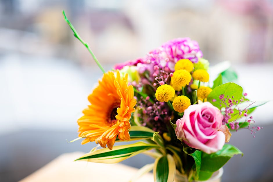 A vibrant floral bouquet featuring an orange gerbera daisy with large, bright petals and a dark center, pink roses with delicate, tightly curled petals, and small yellow billy balls (Craspedia) arrang