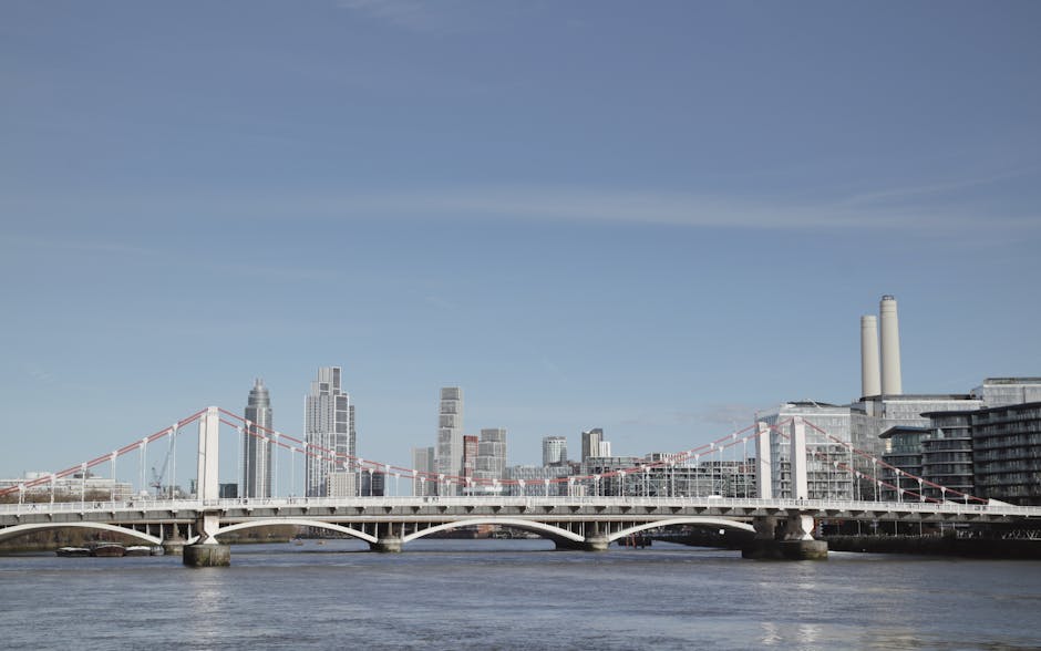 A daytime view of the London skyline featuring the Vauxhall Bridge spanning the River Thames, with modern high-rise buildings and office towers in the background, under a clear blue sky. The bridge ha